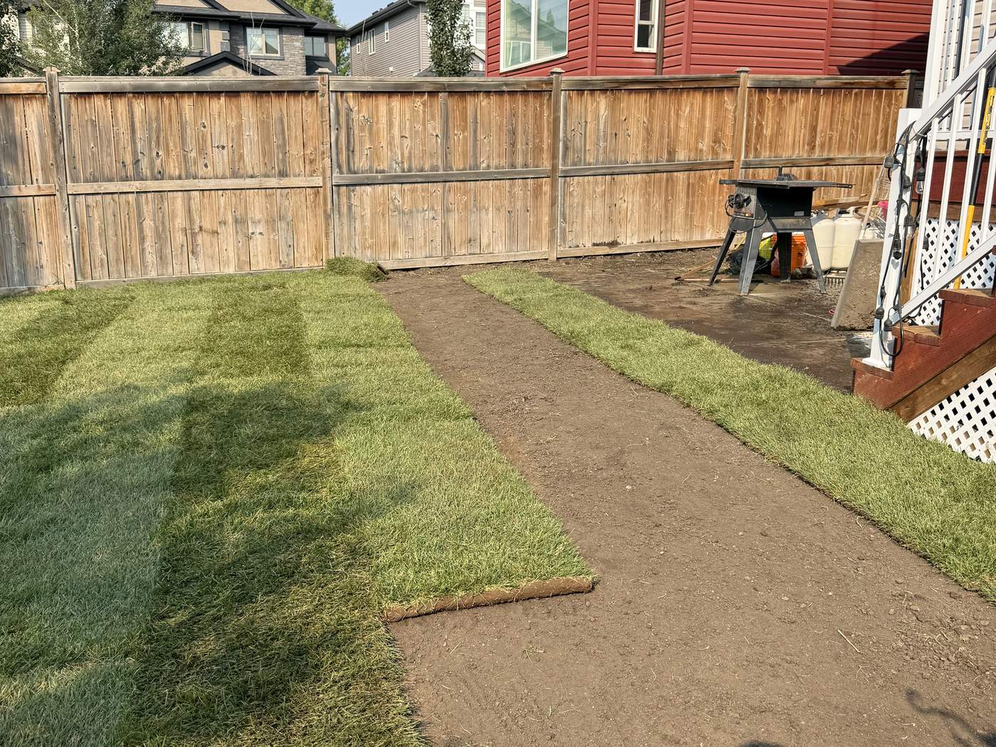 New sod being laid in rows across the Sage Hill back yard, bare graded soil still showing between the rows, the deck stairs at the right