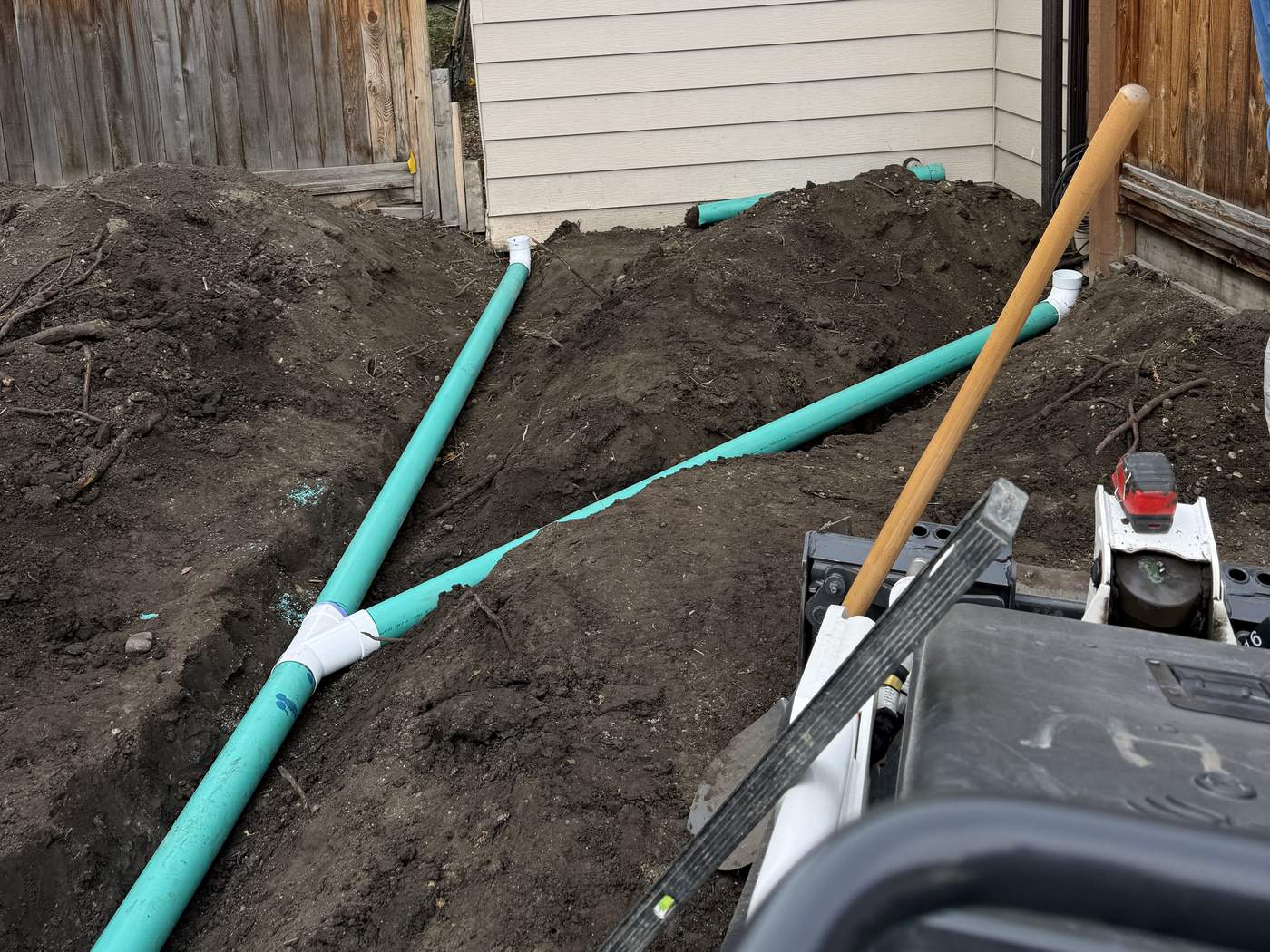 Two green SDR 35 PVC downspout lines joined at a white wye fitting in an open trench beside the house