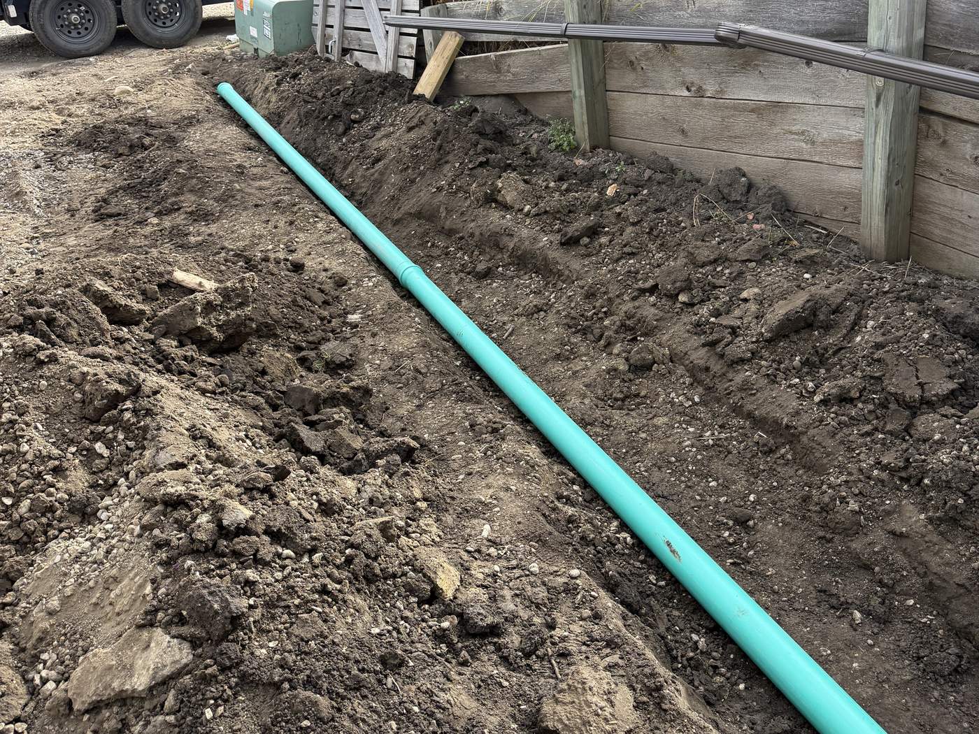 A straight run of green SDR 35 PVC pipe laid in a trench along a wooden retaining wall