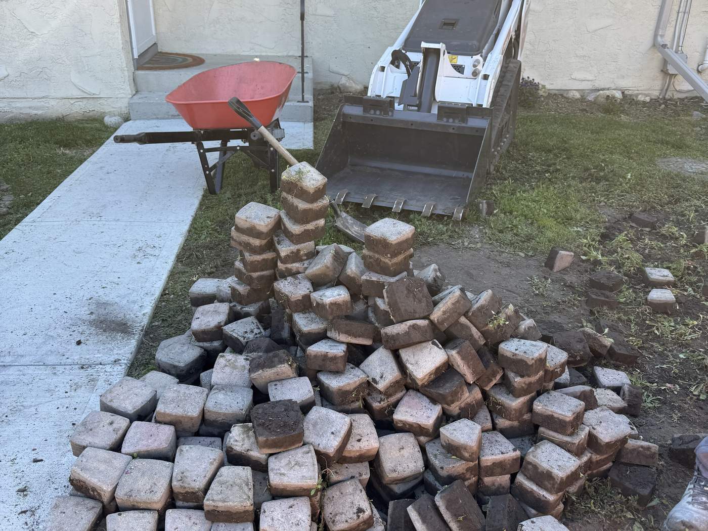 A pile of pavers lifted from the old paver circle, with a wheelbarrow and our Bobcat MT100 bucket behind