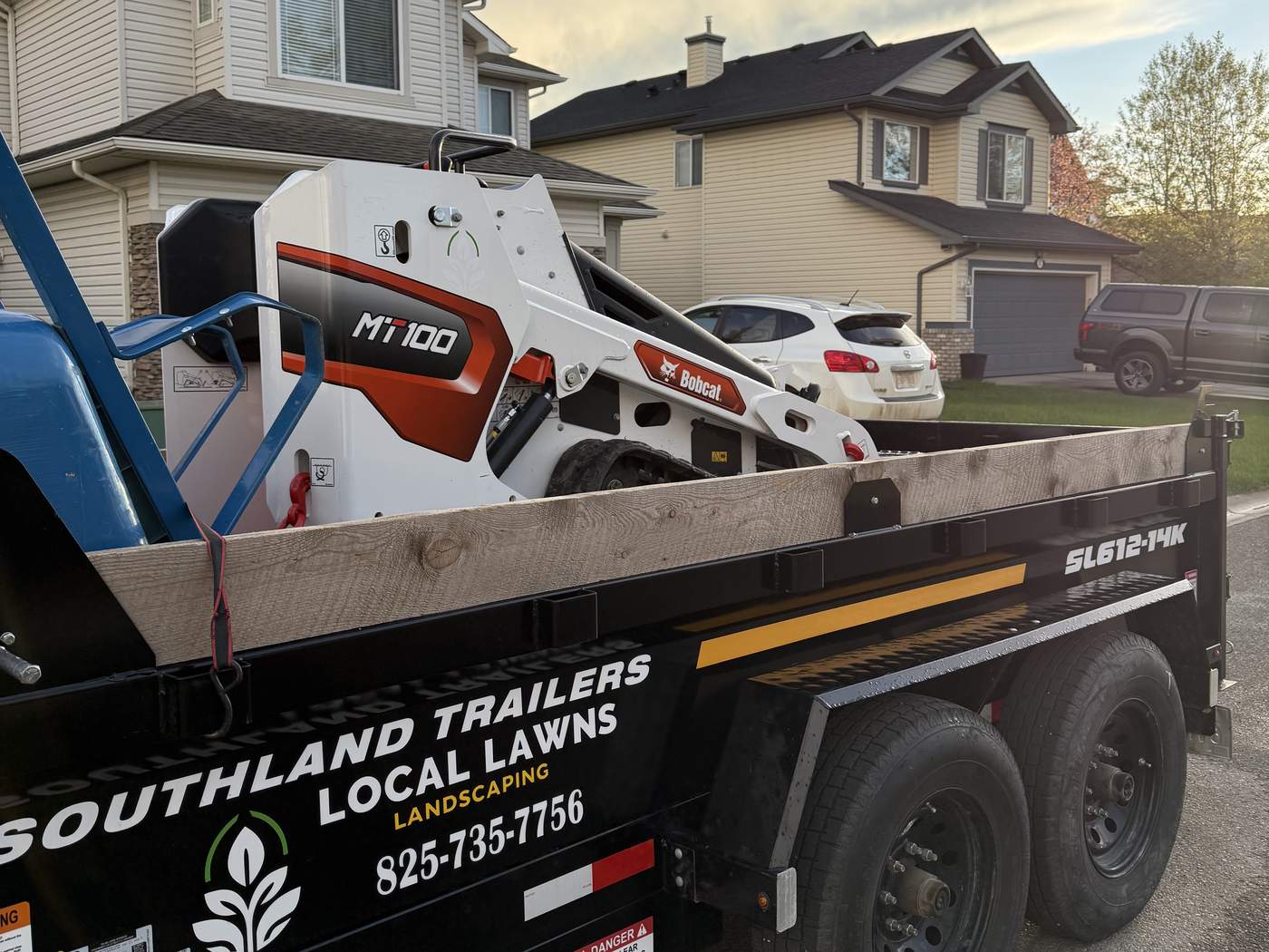 The Local Lawns Bobcat MT100 track loader strapped down on the company dump trailer, lettered Local Lawns Landscaping 825-735-7756, parked on a residential street