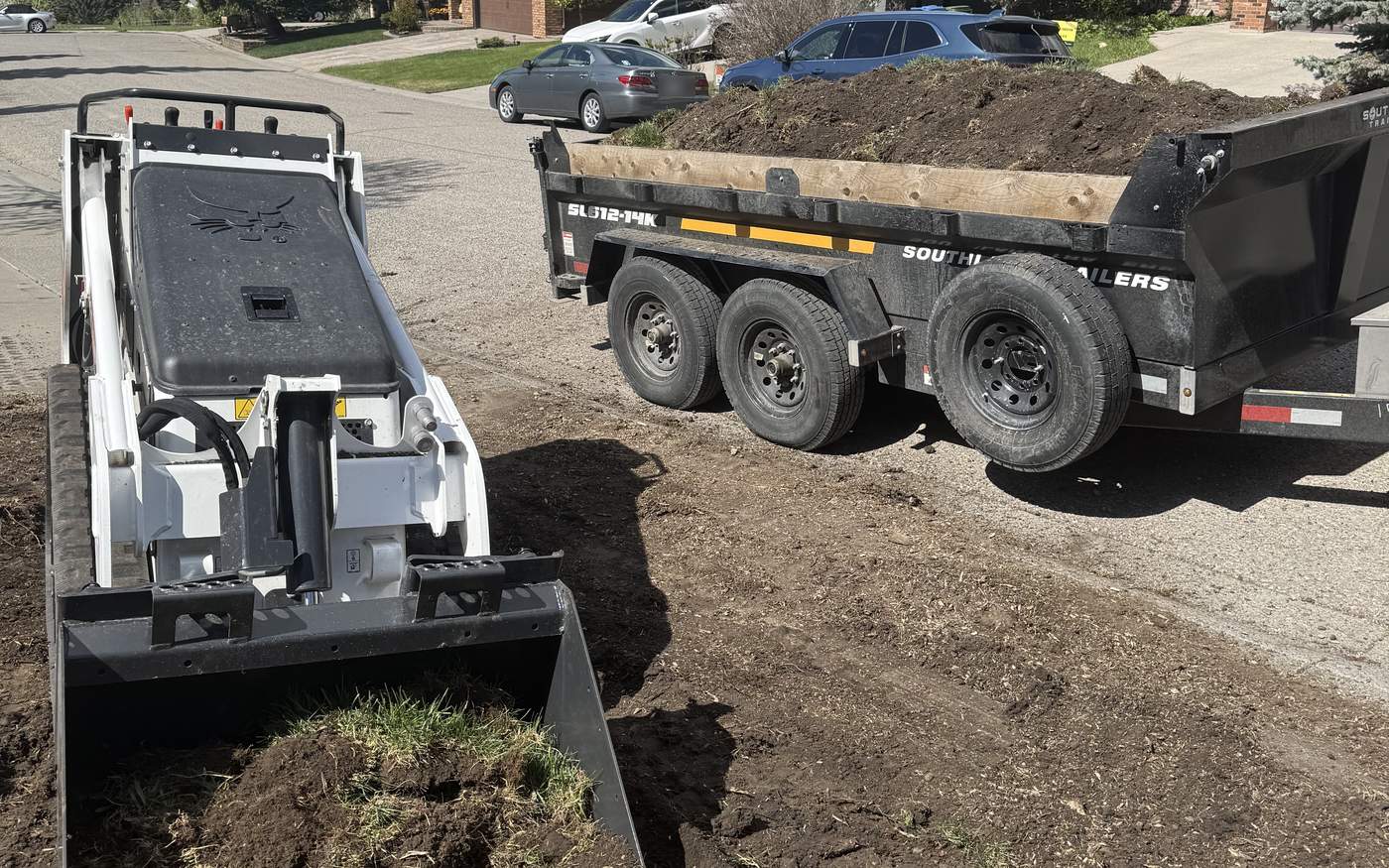 Bobcat track loader holding a bucket of stripped sod on a Calgary sod replacement job, beside a dump trailer heaped with torn-out turf and soil