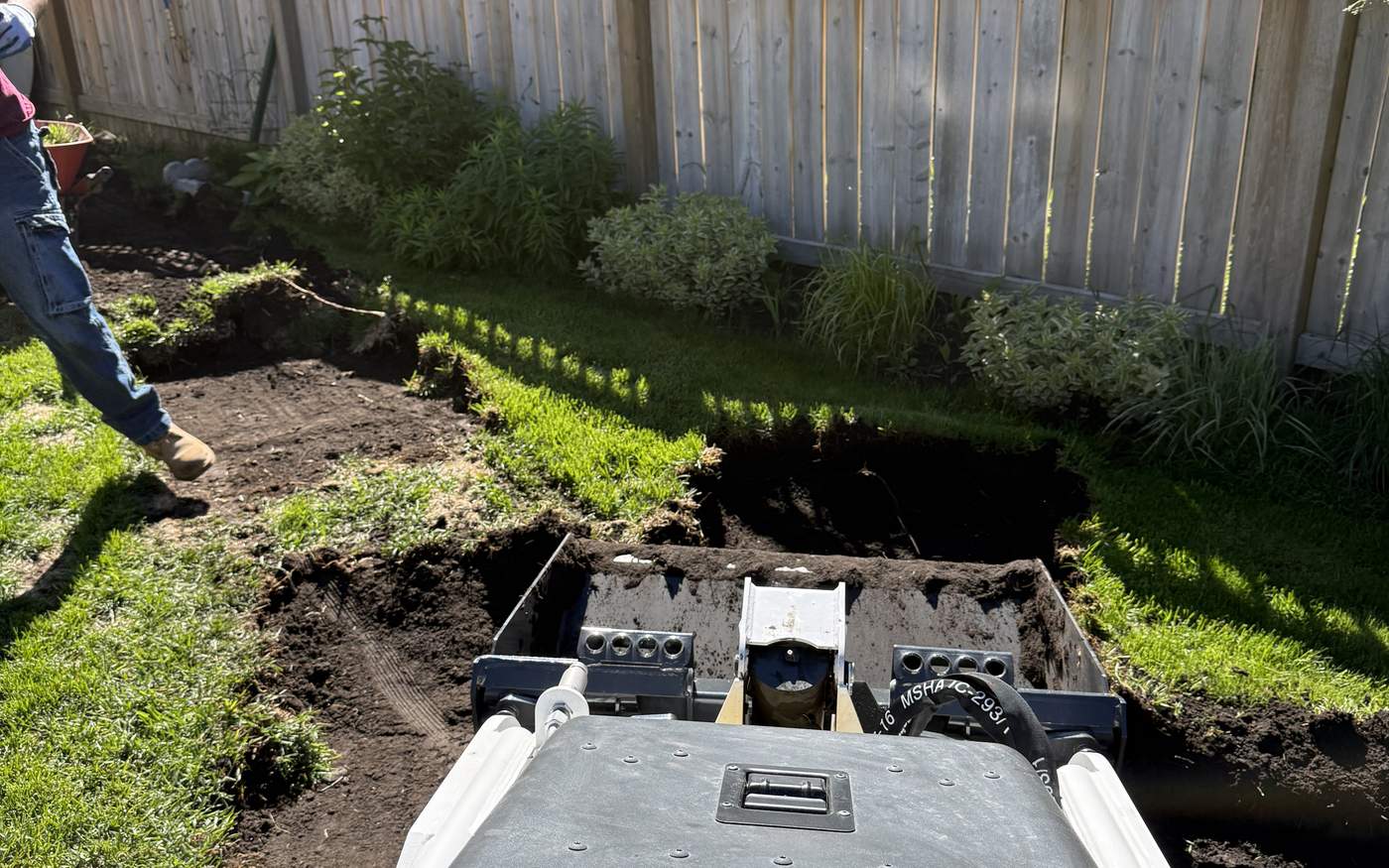 Mini excavator cutting a French drain trench along a fence line in a Calgary yard, mid-dig
